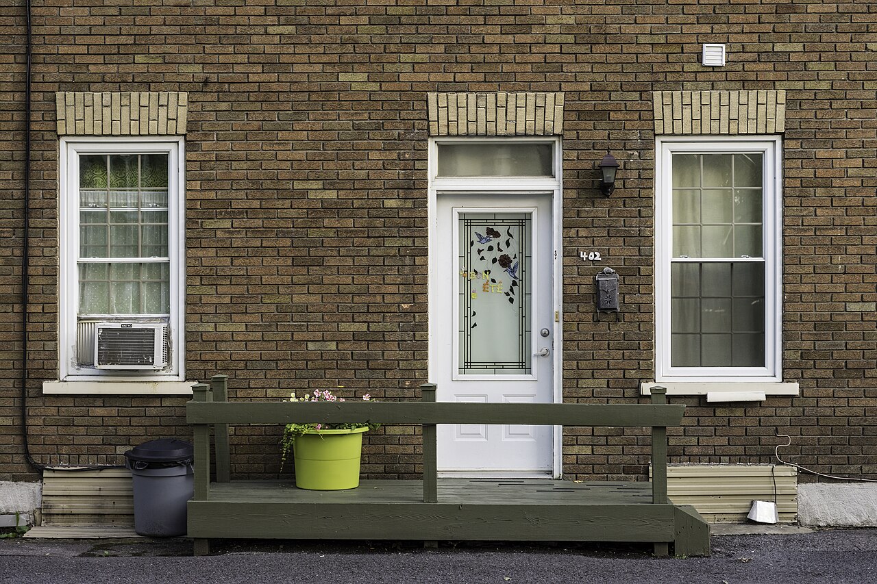 A low green wooden porch with a pot of flowers runs along a brown brick house front with a white door.