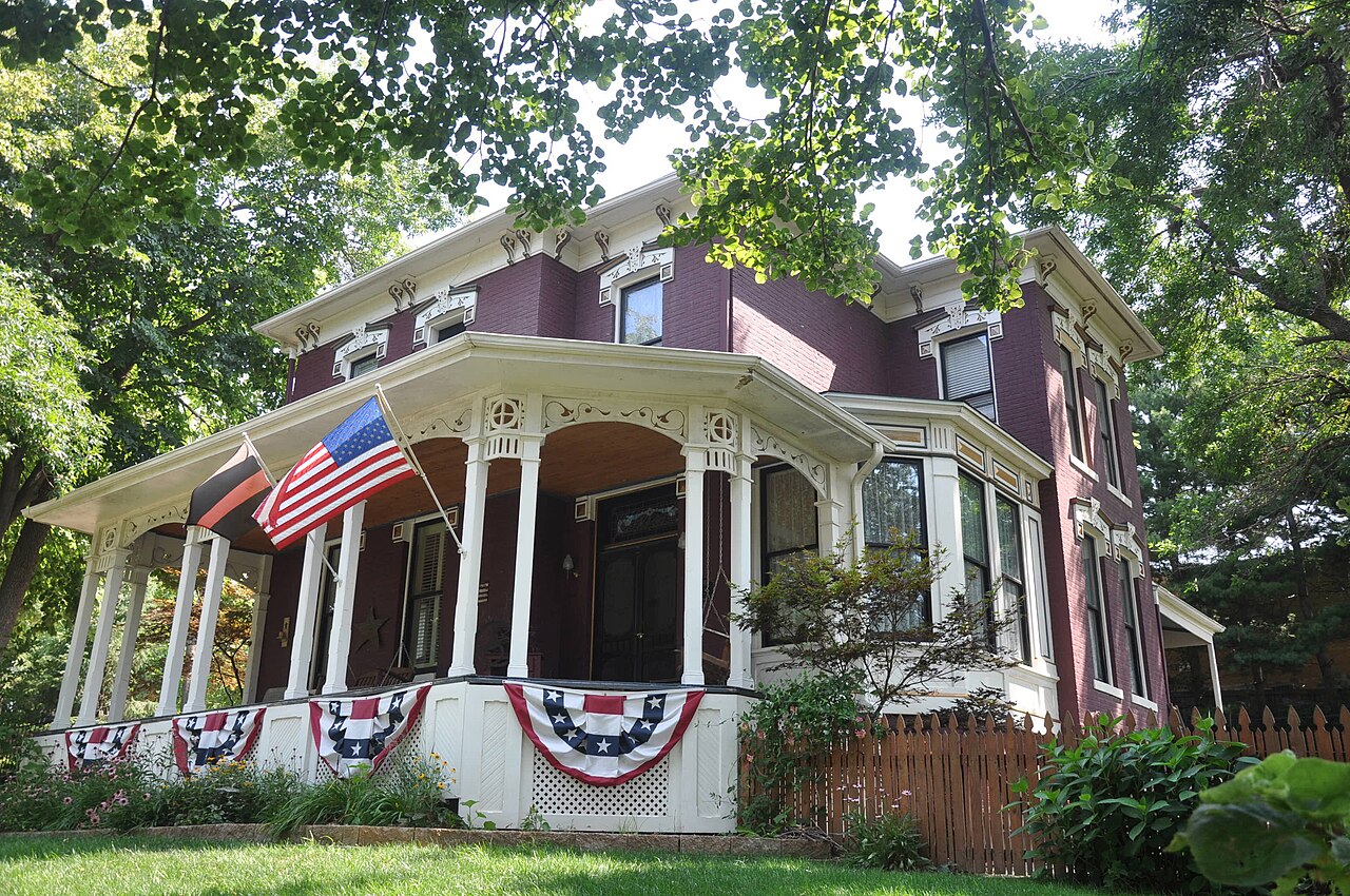A dark red Victorian house with a white wraparound porch hung with flag bunting, shaded by large trees.