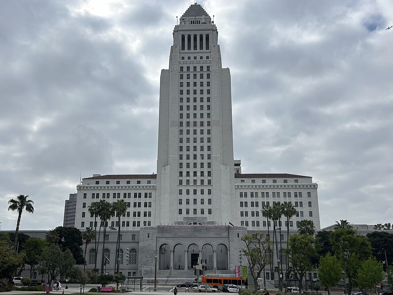 A white stepped tower rises above a wide columned base, with palm trees in front and a cloudy sky behind.