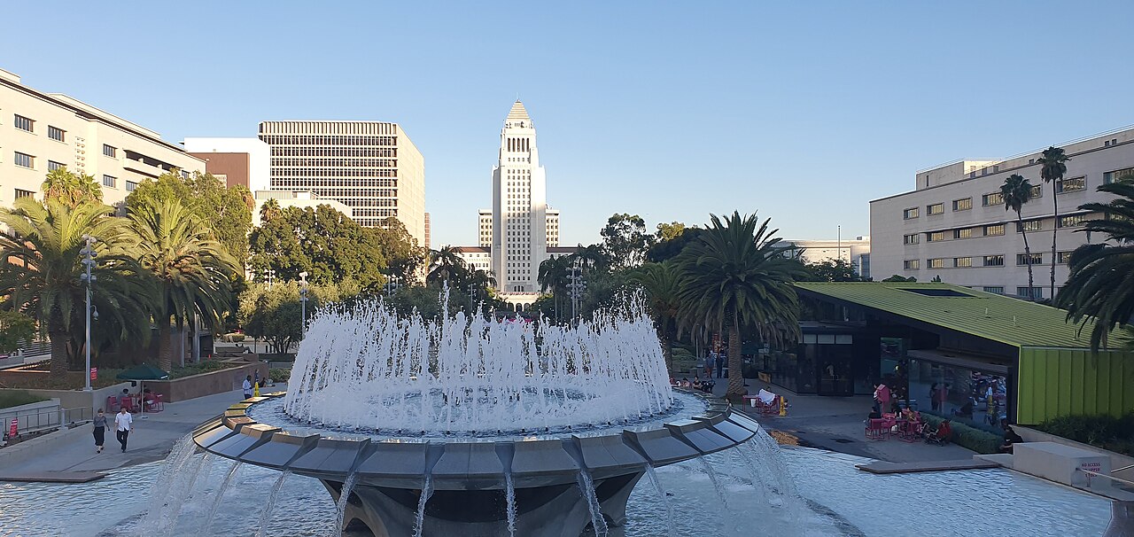 A circular fountain sprays in the foreground, with palm trees and a white tower in the distance under a clear sky.
