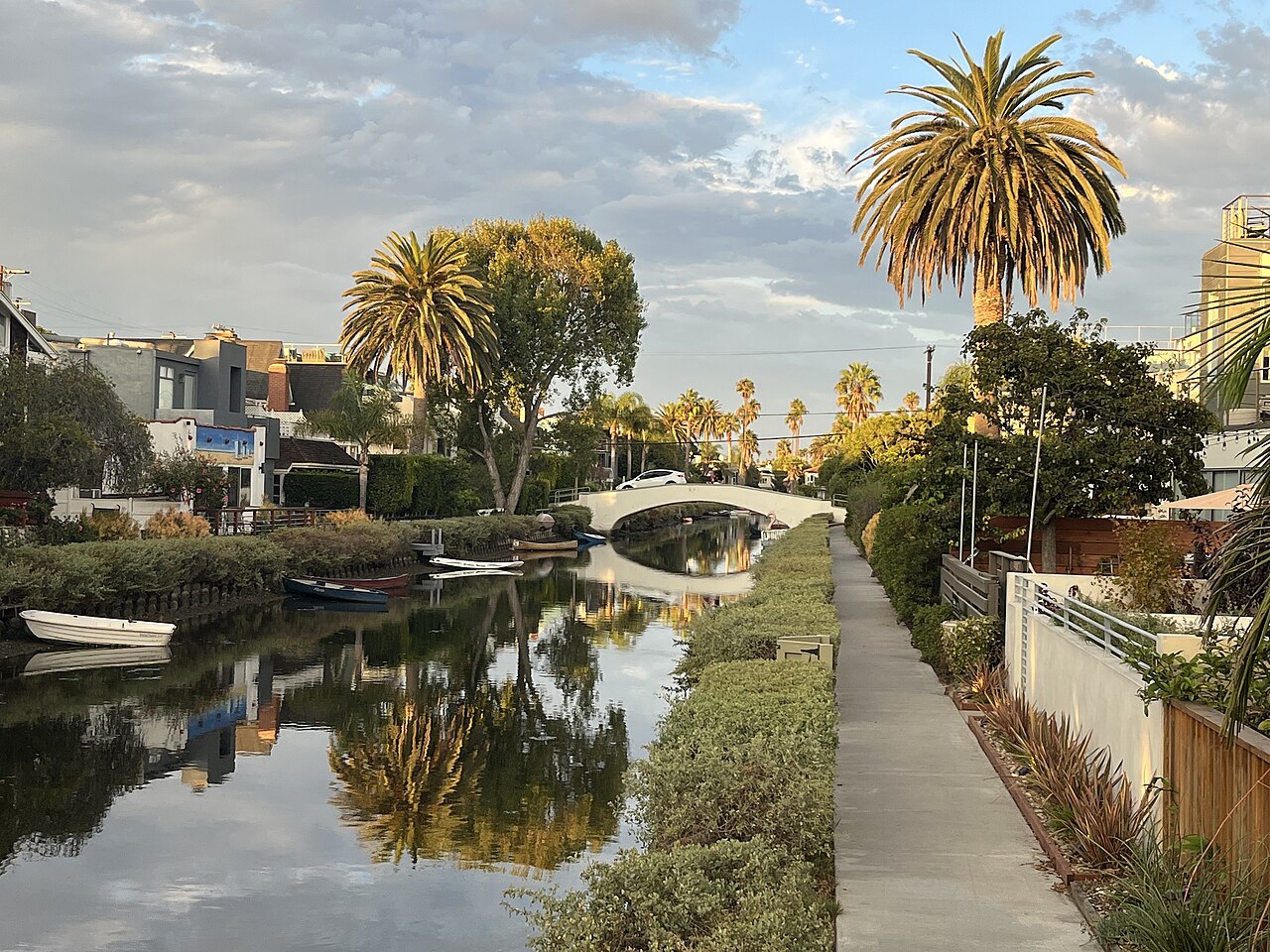 A still canal reflects palm trees and a white arched footbridge, with a paved path and garden hedges alongside.