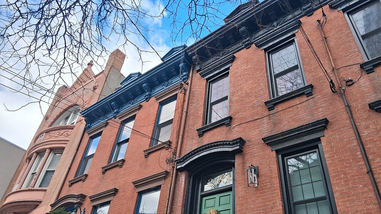 A row of red brick town houses with dark cornices and tall windows, seen from the pavement looking up.