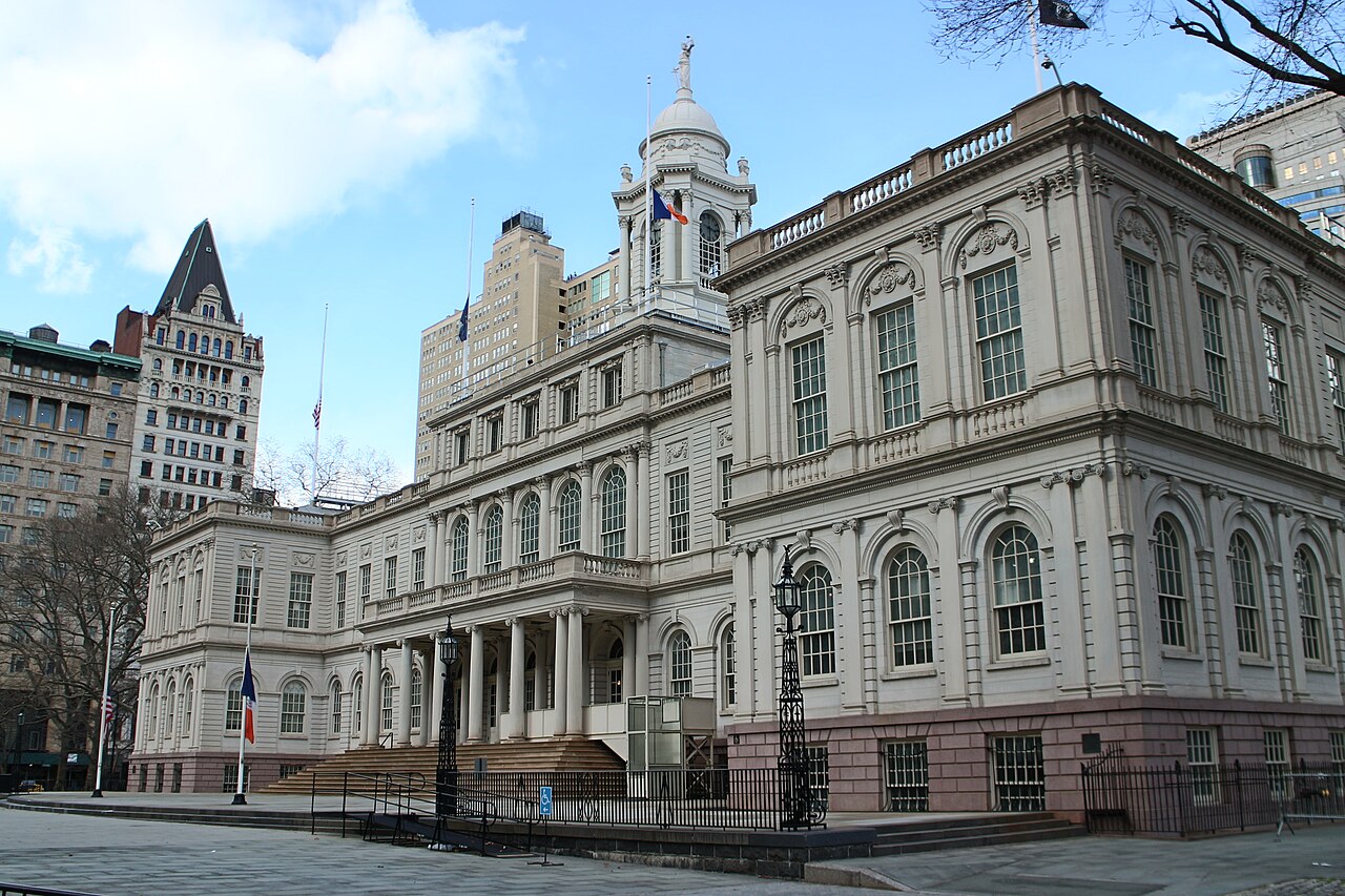 A pale stone civic building with arched windows and a central cupola stands behind bare trees under a blue sky.