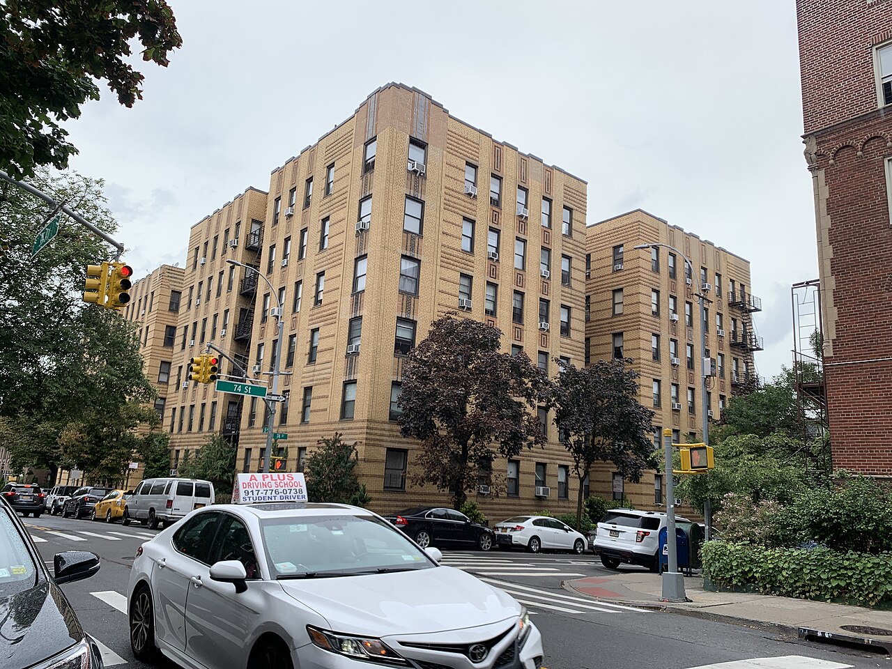 Six-storey tan brick apartment blocks stand on a street corner with parked cars and street trees.