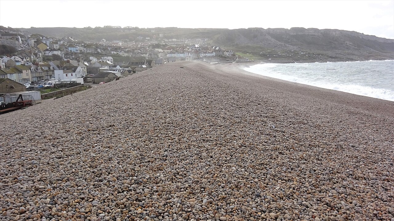 A broad bank of brown pebbles curves towards a hillside village, with white surf on the right.