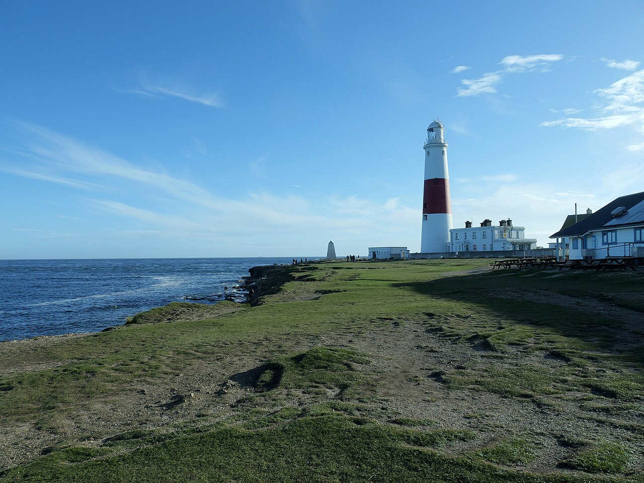 A red and white striped lighthouse stands on a grassy headland beside low white buildings, with the sea to the left.