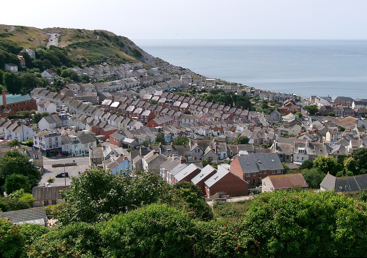 Rows of terraced houses step down a green hillside towards a calm grey sea.