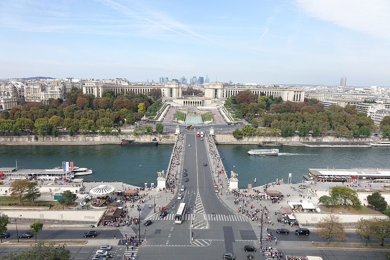 A bridge crosses a river towards formal gardens and a curved palace, with the city spreading out behind.