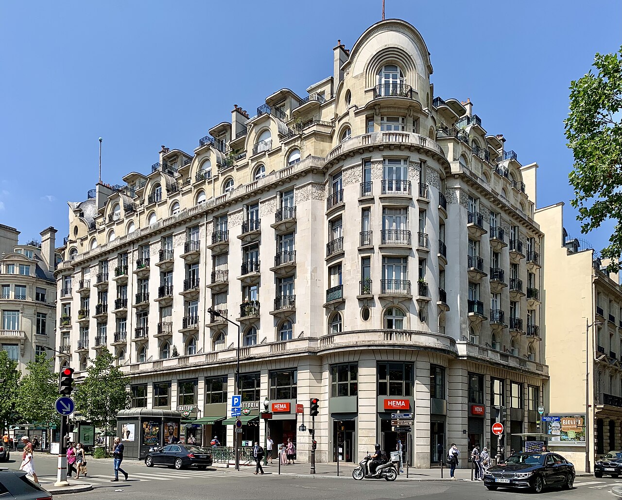 A cream stone corner building with rounded bays, iron balconies and a domed roof stands over a street junction.