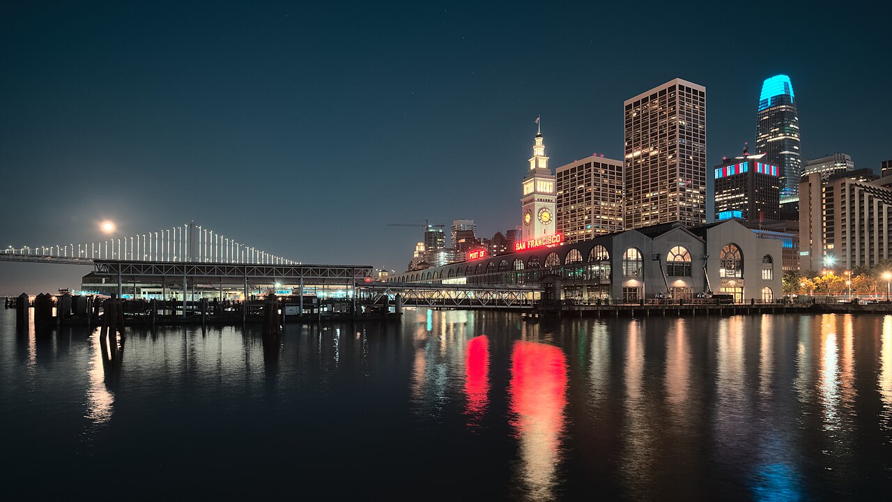 A lit clock tower and office towers reflect in dark water, with a suspension bridge and the moon to the left.