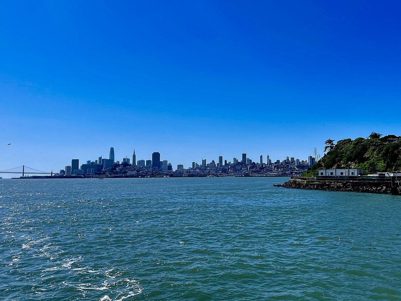 A line of towers sits on the far shore of blue water, with a wooded point on the right.