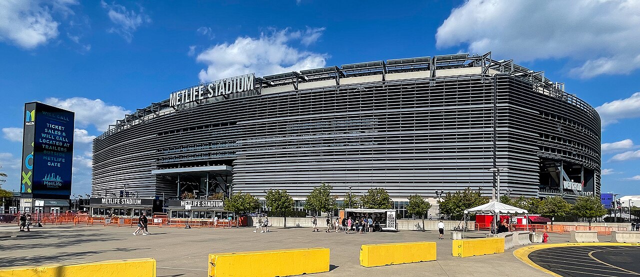 A large oval stadium clad in grey horizontal louvres stands behind a paved forecourt under a blue sky.