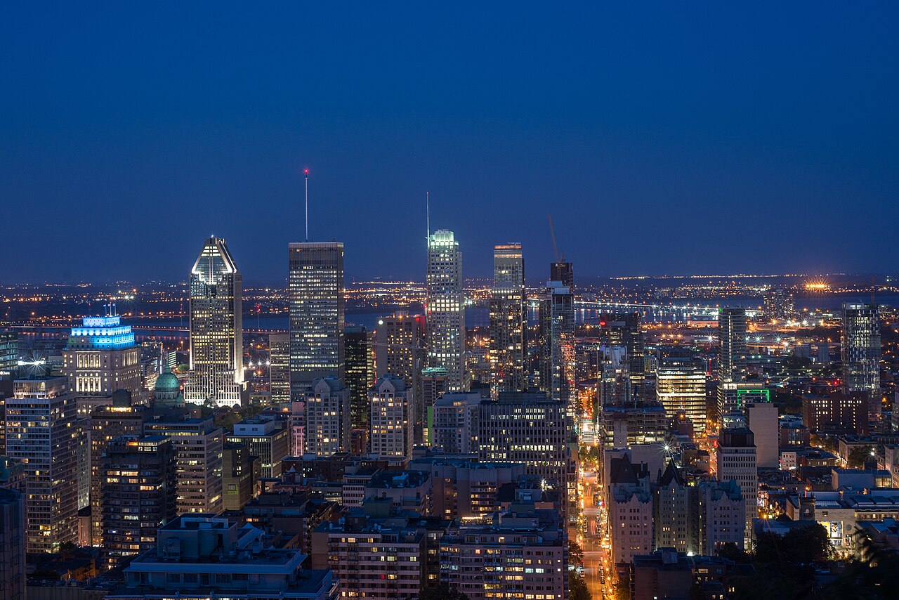 Lit office towers stand against a deep blue evening sky, with city lights stretching to the horizon.