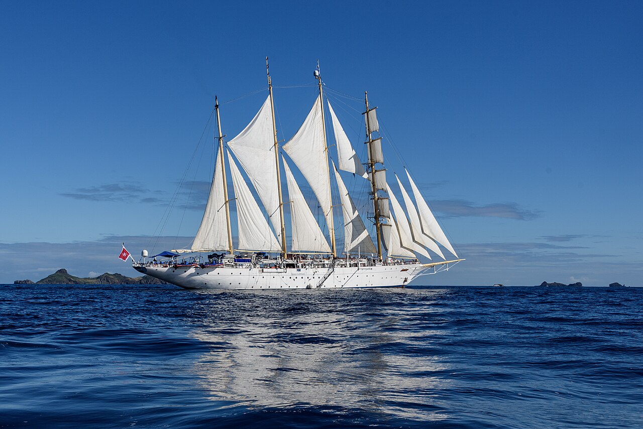 A white tall ship under full sail crosses deep blue water, with small rocky islands on the horizon.