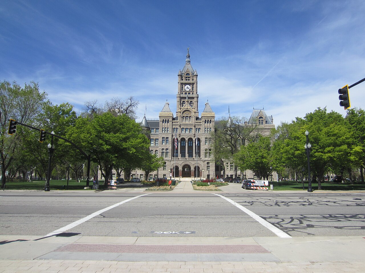 A grey stone building with a tall central clock tower stands behind green trees across an empty road junction.