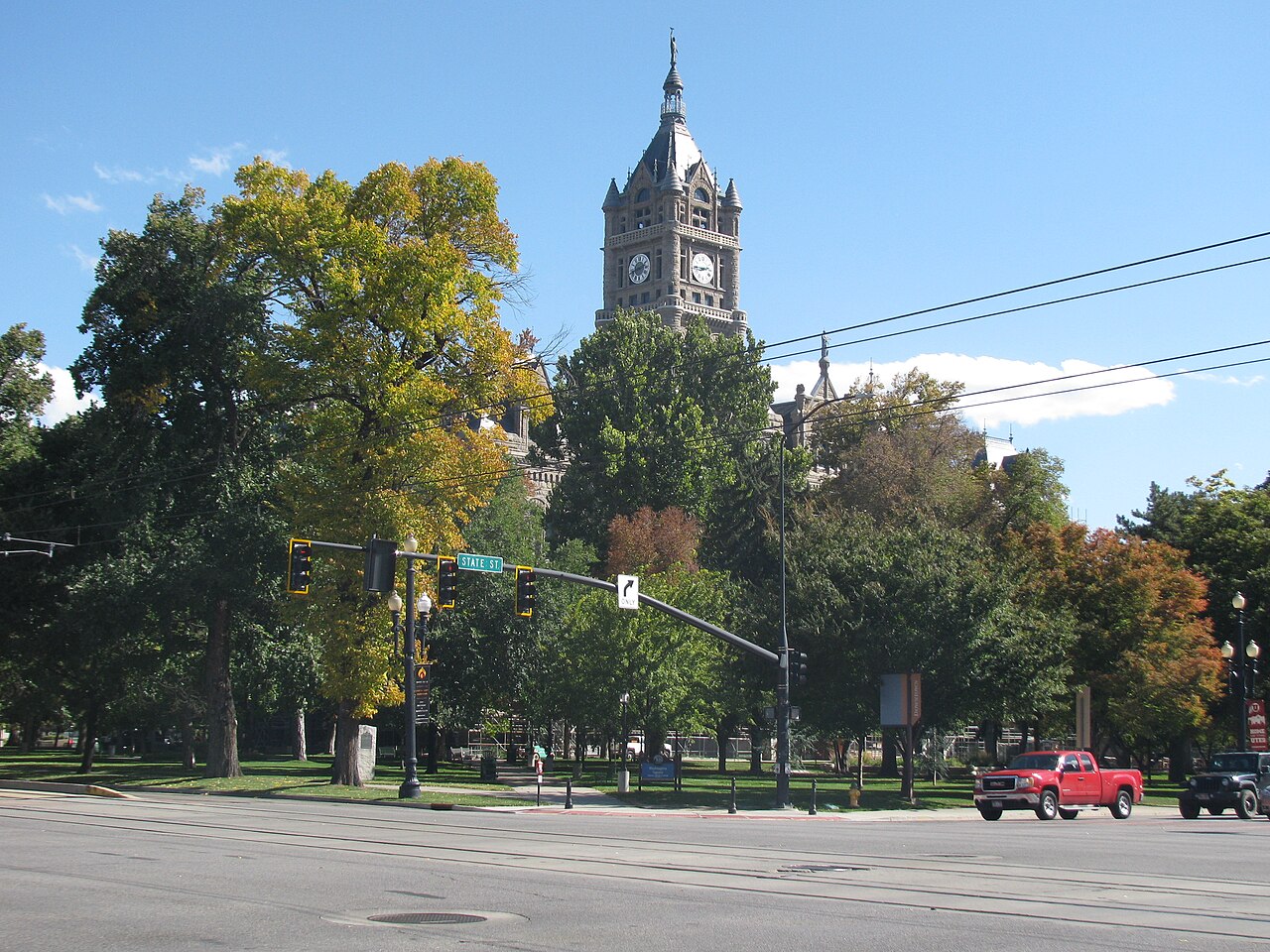 A stone clock tower rises above autumn trees beside a road with a red pickup truck.