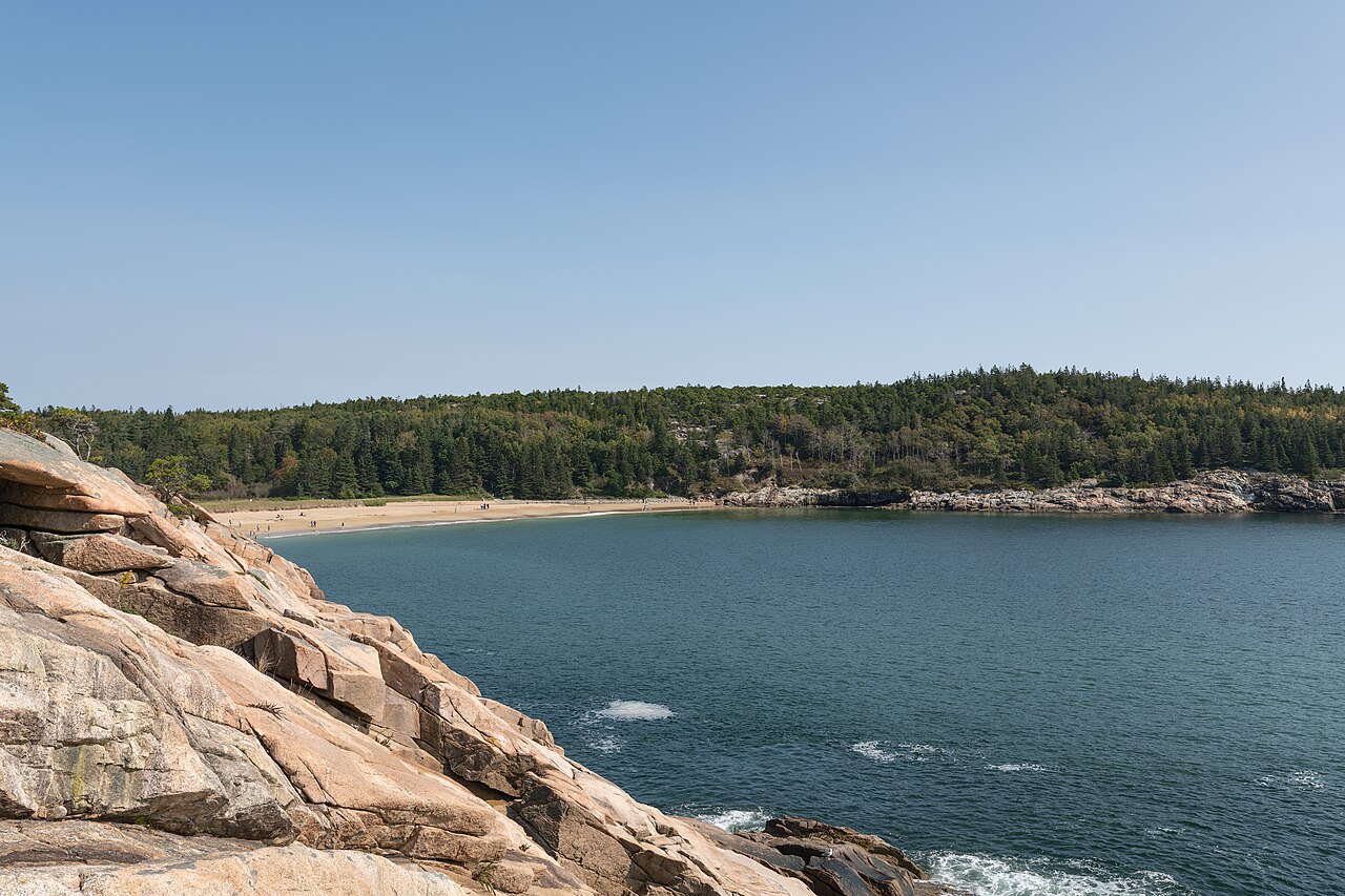 Pink granite rocks slope into a blue cove, with a sandy beach and a forested hill on the far side.