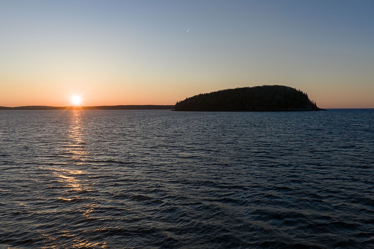The sun sits low on the horizon beside a small wooded island, its light reflected across calm water.