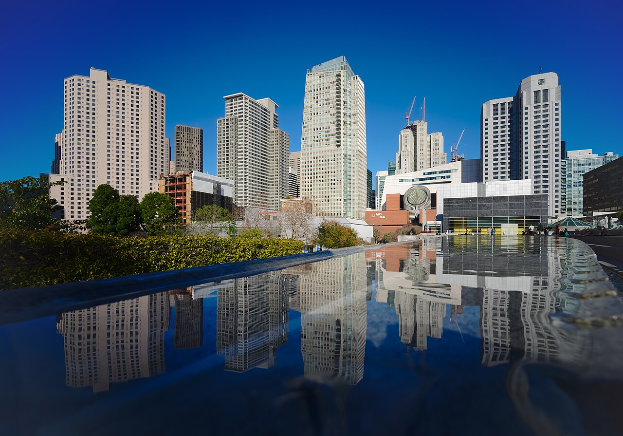Pale high-rise towers under a deep blue sky are mirrored in the still surface of a pool.