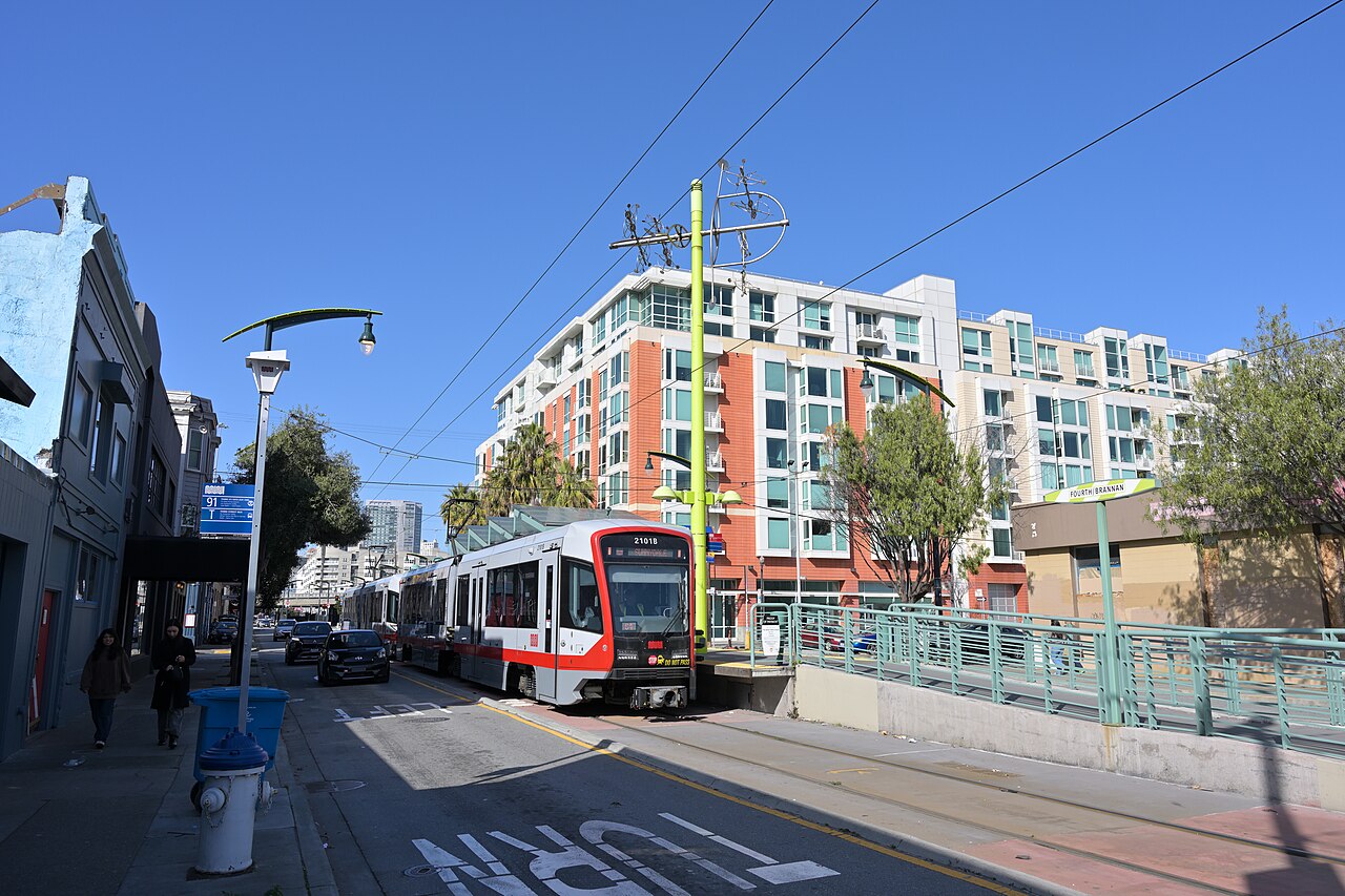 A red and silver light-rail train waits at a street platform, with a new apartment block behind it.