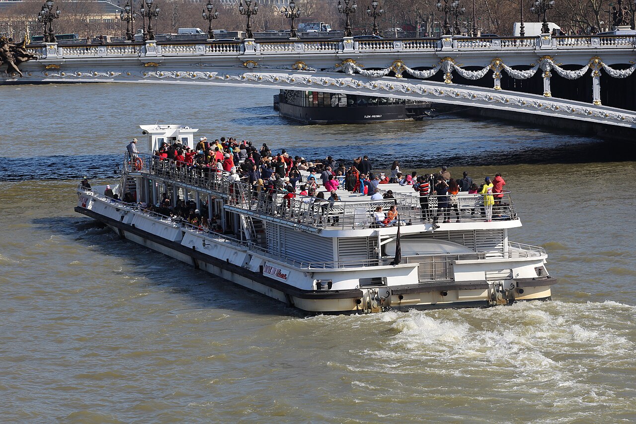 An open-decked tour boat full of passengers moves along a river towards a low ornate bridge.