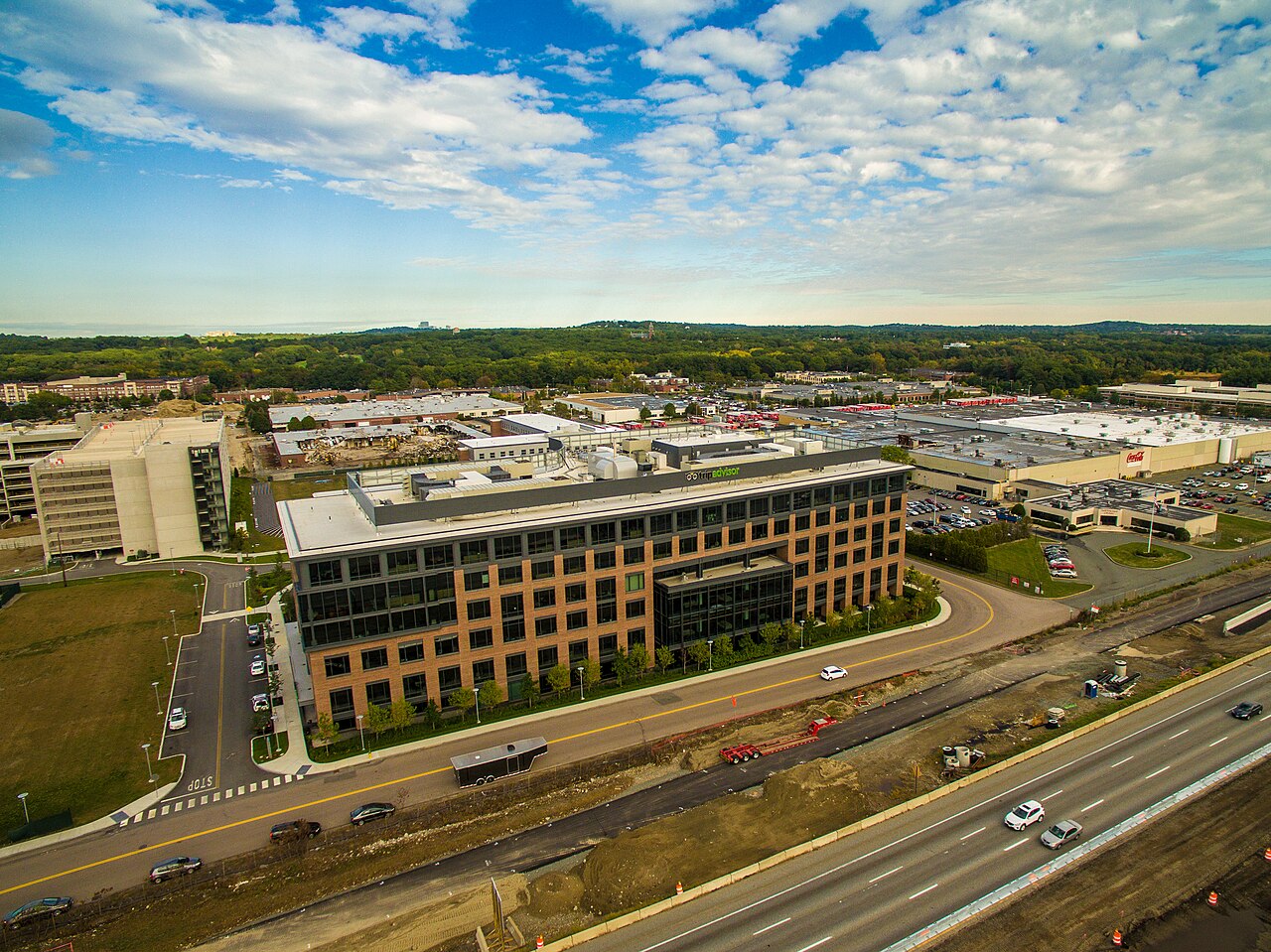 A long brick and glass office building sits beside a highway, with car parks and woodland around it.