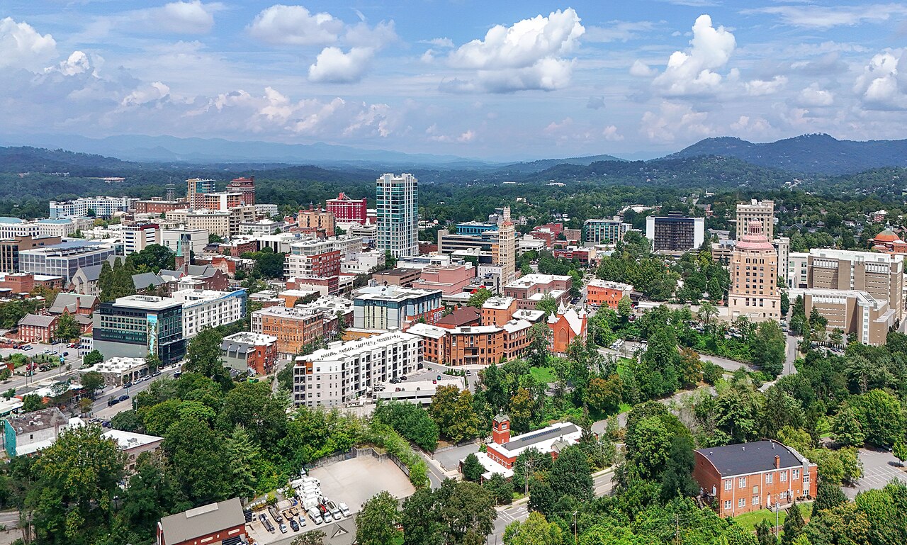 A compact downtown of brick and concrete buildings is surrounded by trees, with blue mountain ridges behind.