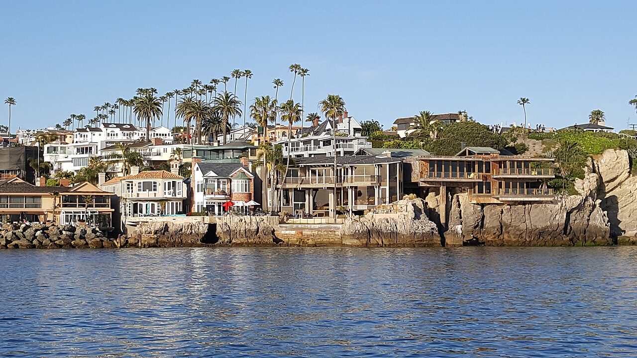 Large houses and palm trees crowd a rocky shoreline above calm blue water.