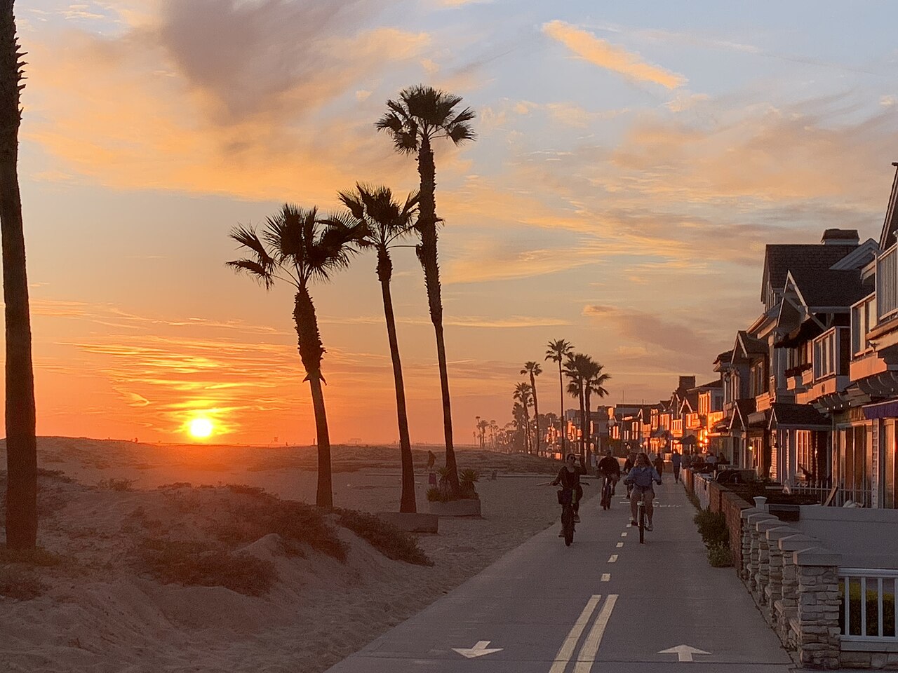 Palm trees stand in silhouette against an orange sunset beside a paved path lined with beach houses.