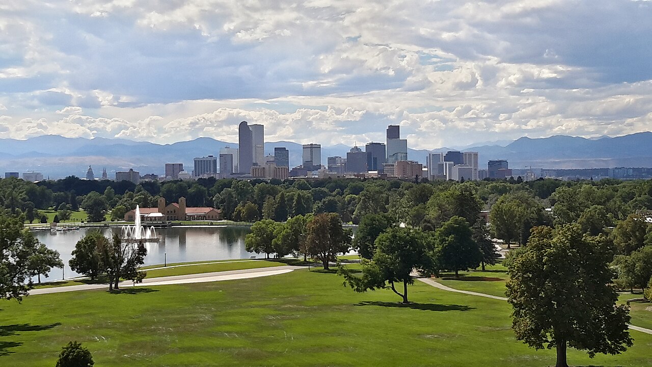A cluster of office towers rises beyond a park lake and green lawns, with mountains on the horizon.