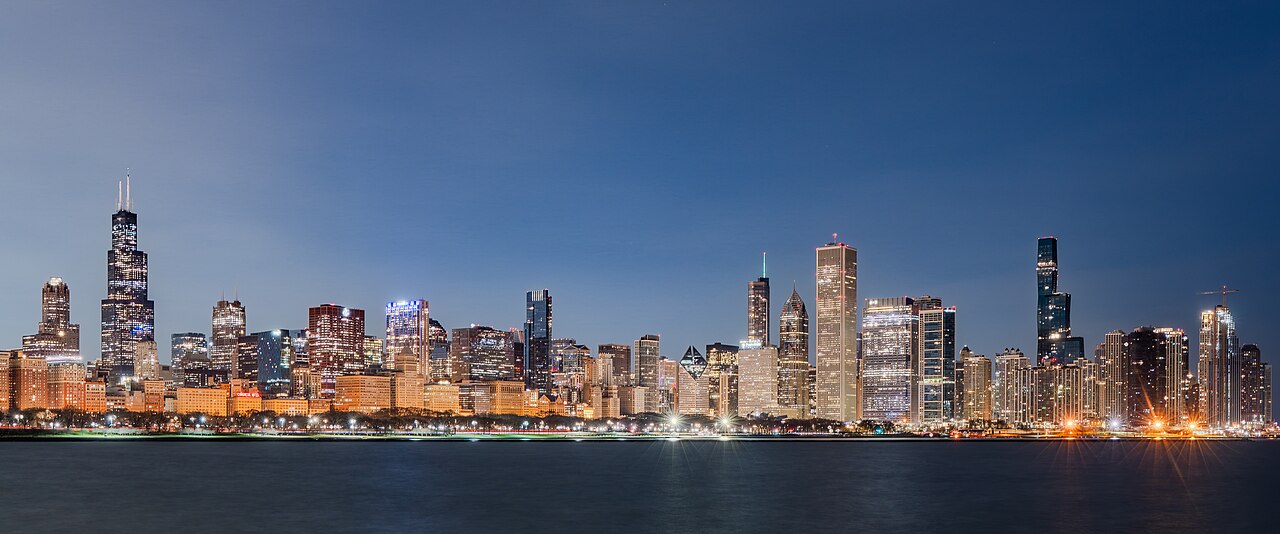 Lit skyscrapers line the far shore of a dark lake under a deep blue dawn sky.