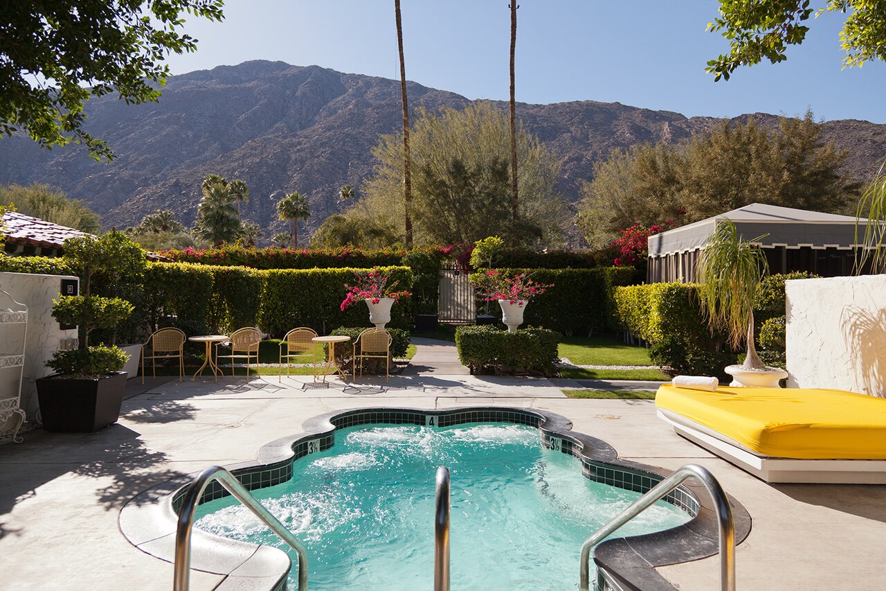 A curved turquoise pool with handrails sits in a hedged garden, with dry mountains rising behind.