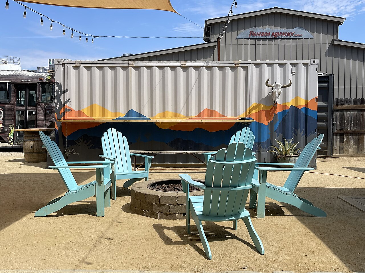 Four turquoise wooden chairs circle a round stone fire pit on a gravel patio in front of a painted shipping container.