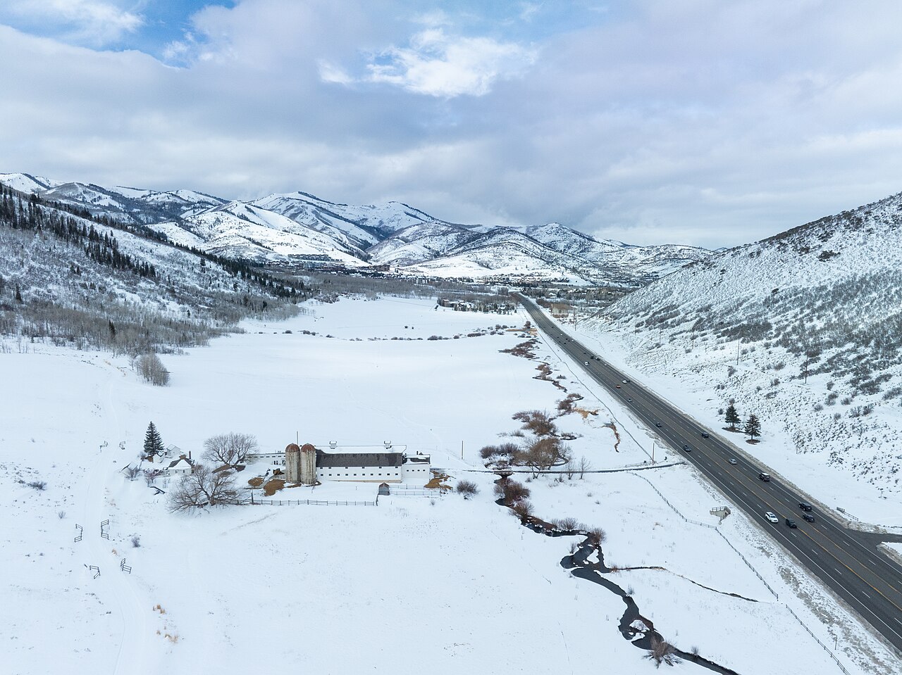 A snow-covered valley with a road, a winding creek and scattered buildings runs toward white mountains.