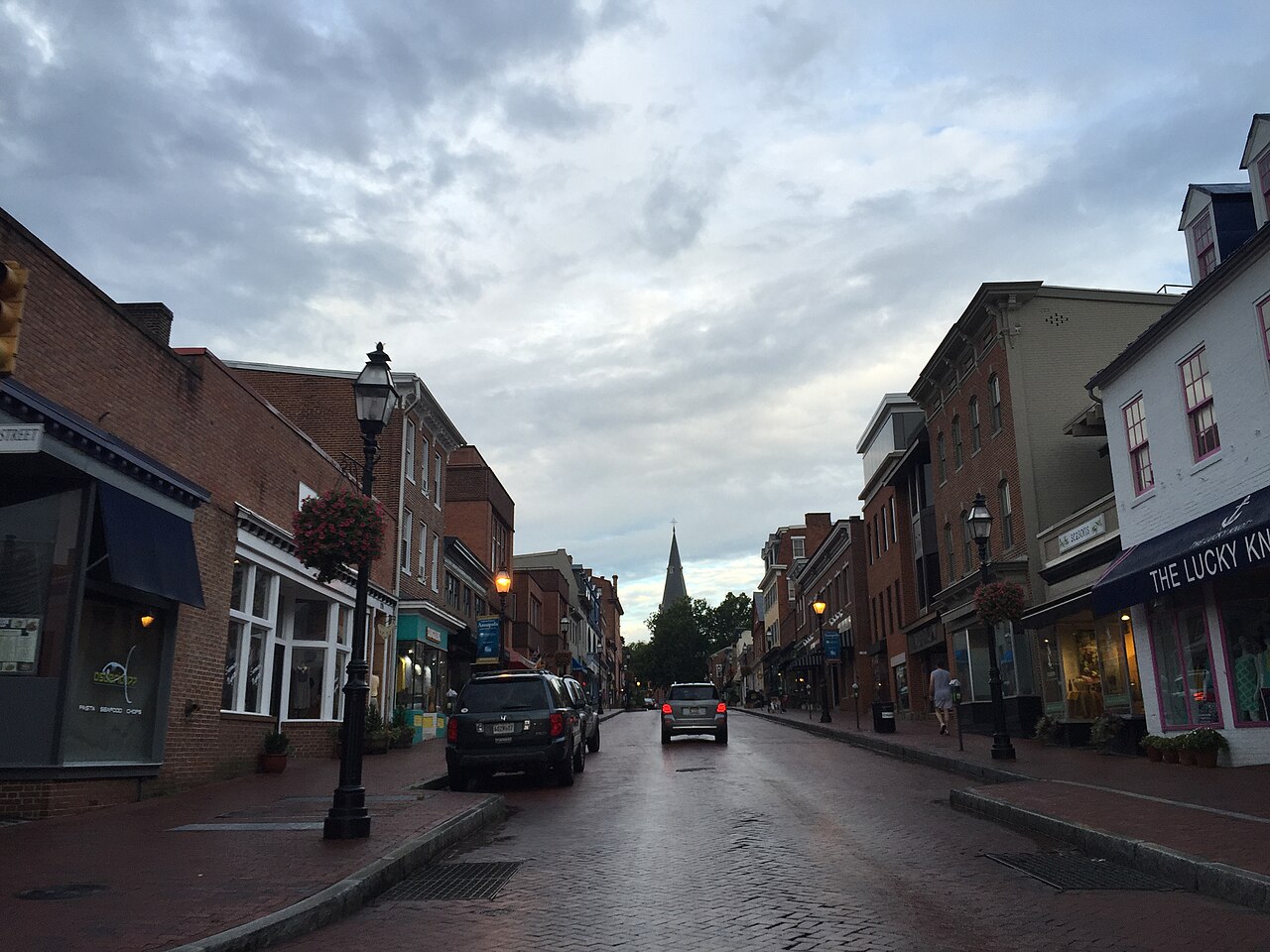 A brick-paved street climbs between old shopfronts toward a church spire under a cloudy sky.