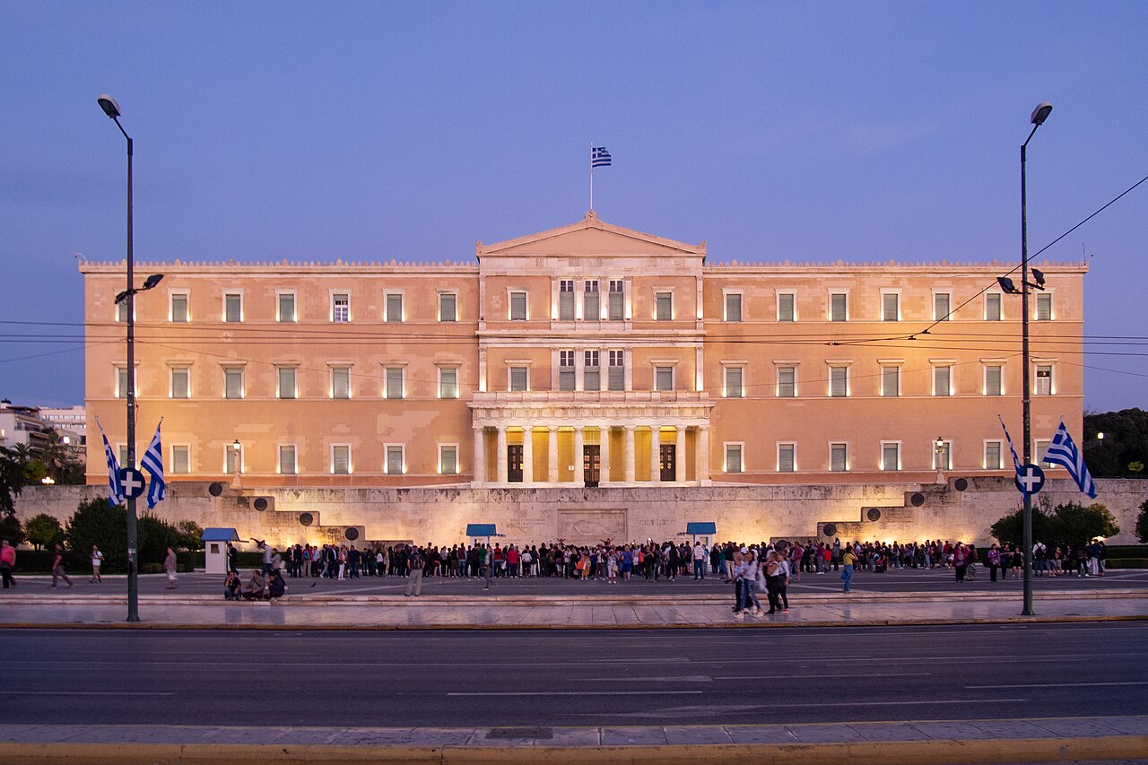 A wide neoclassical building glows pink under floodlights against a violet evening sky.
