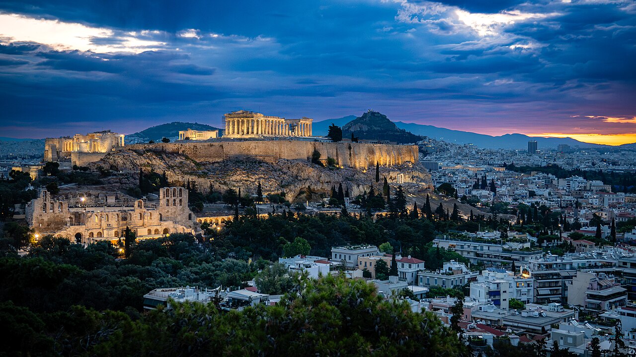 A floodlit ancient temple on a rocky hill stands above a dense city under a dark blue dawn sky.