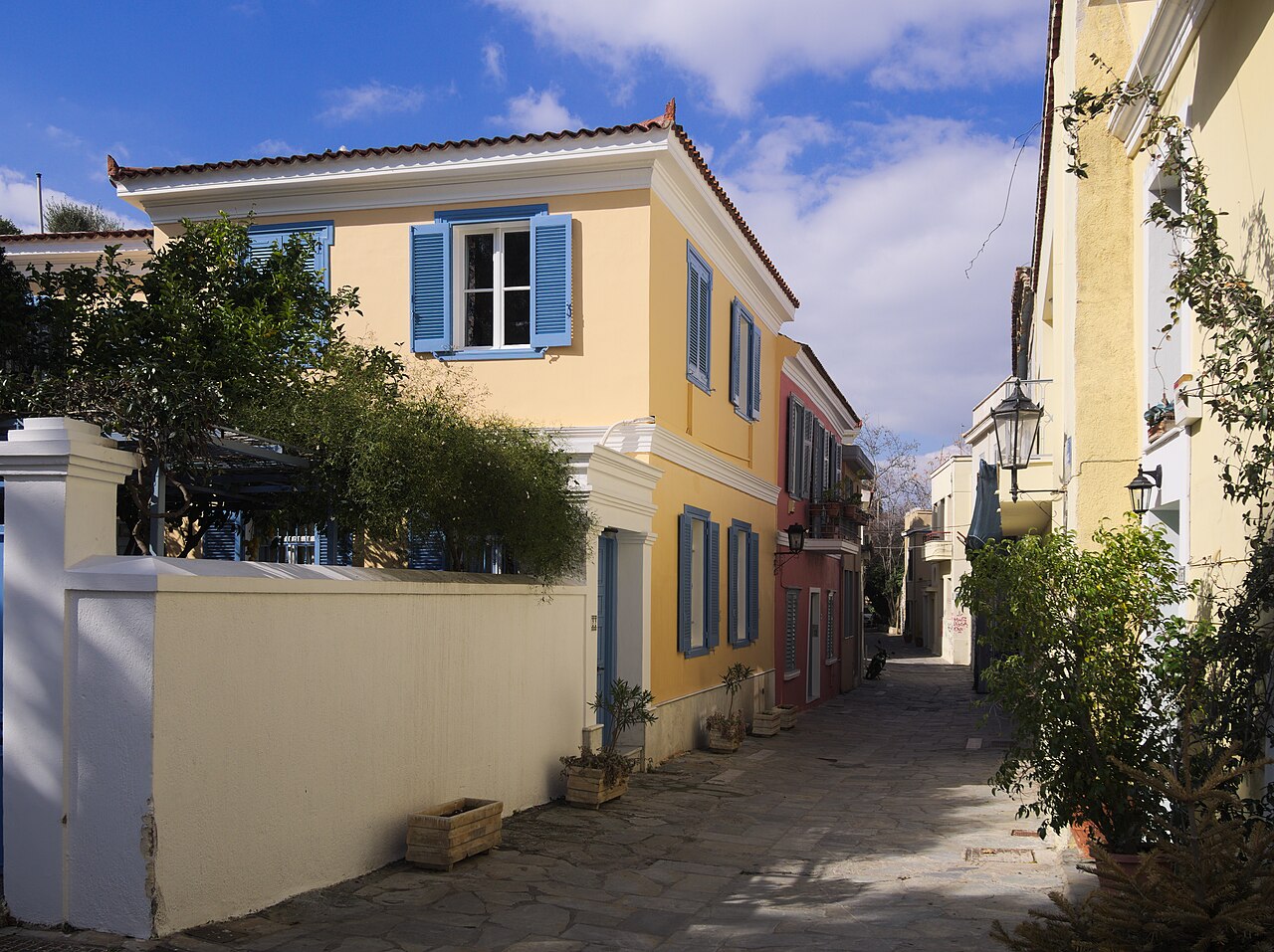 A yellow two-storey house with blue shutters stands beside a narrow lane under a bright sky.