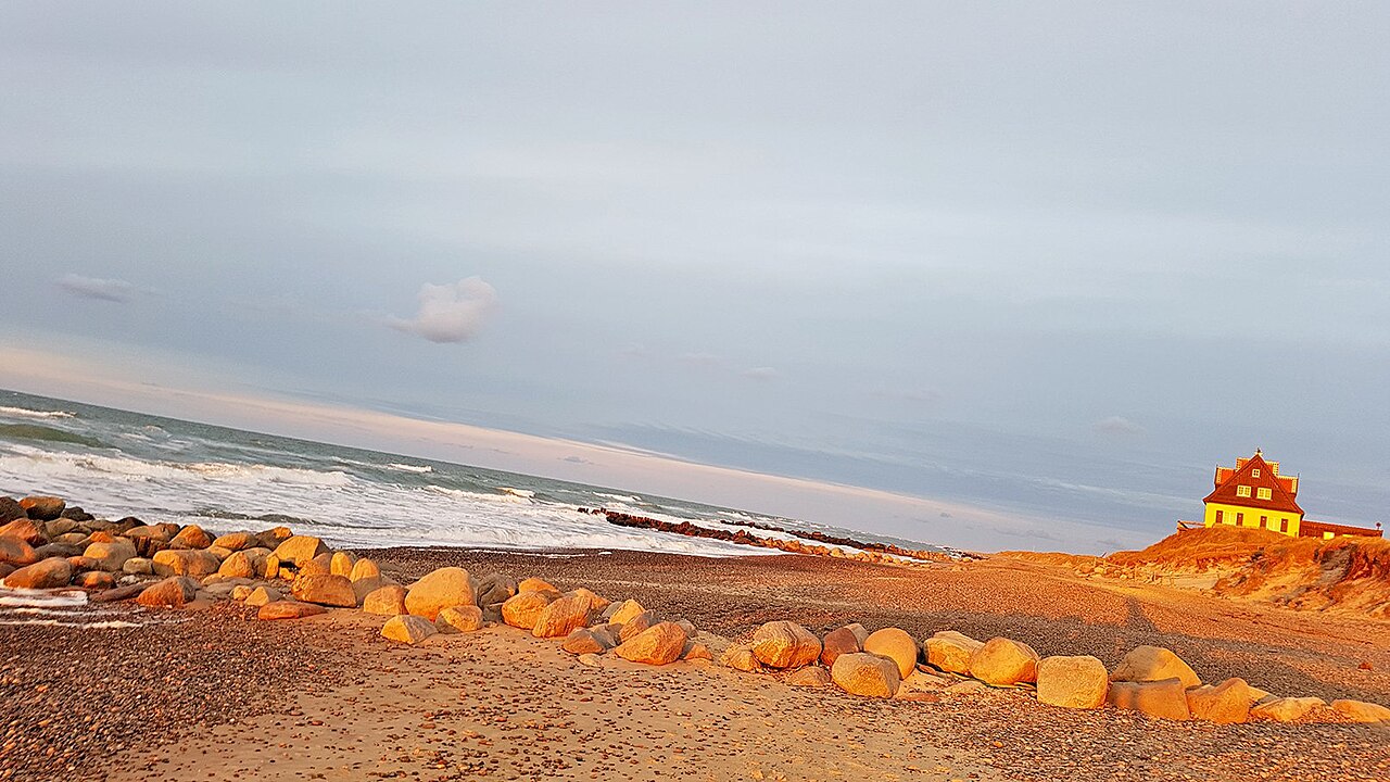 Low golden light falls on sand and a row of boulders beside breaking waves, with a small house at the edge.