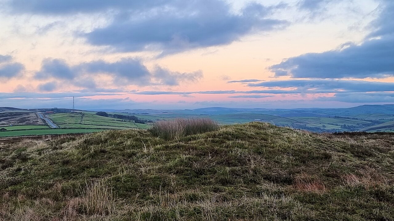 A grassy mound on open moorland looks out over green fields under a pink and blue evening sky.