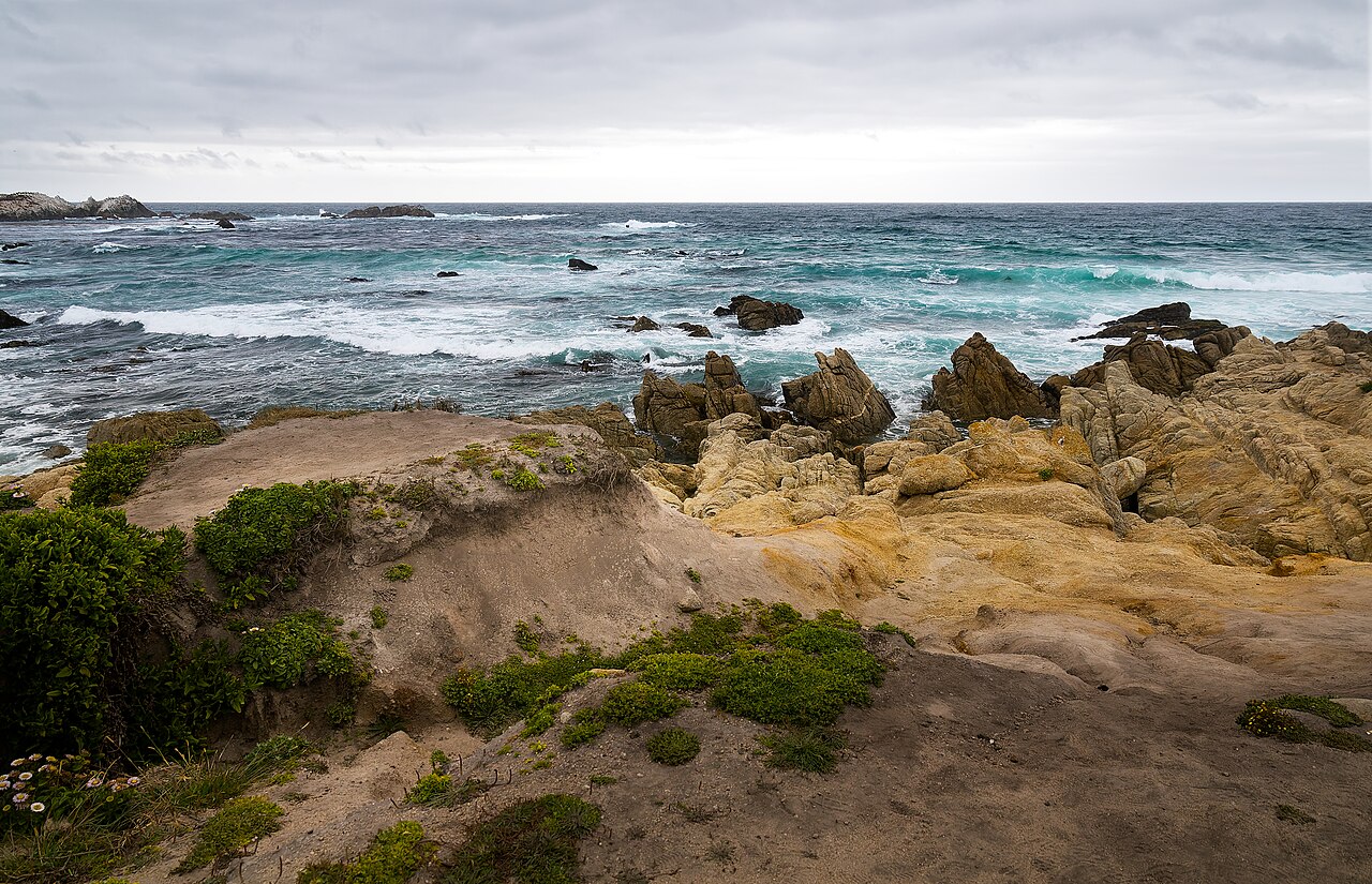 Waves break on a rocky shore below sandy bluffs under a grey sky.
