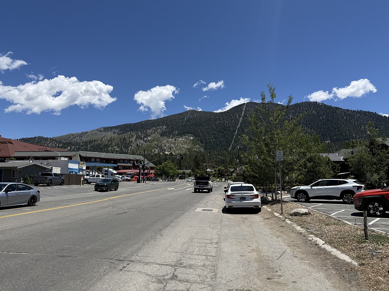 A wide road with parked cars and low buildings runs toward a tree-covered mountain under a blue sky.