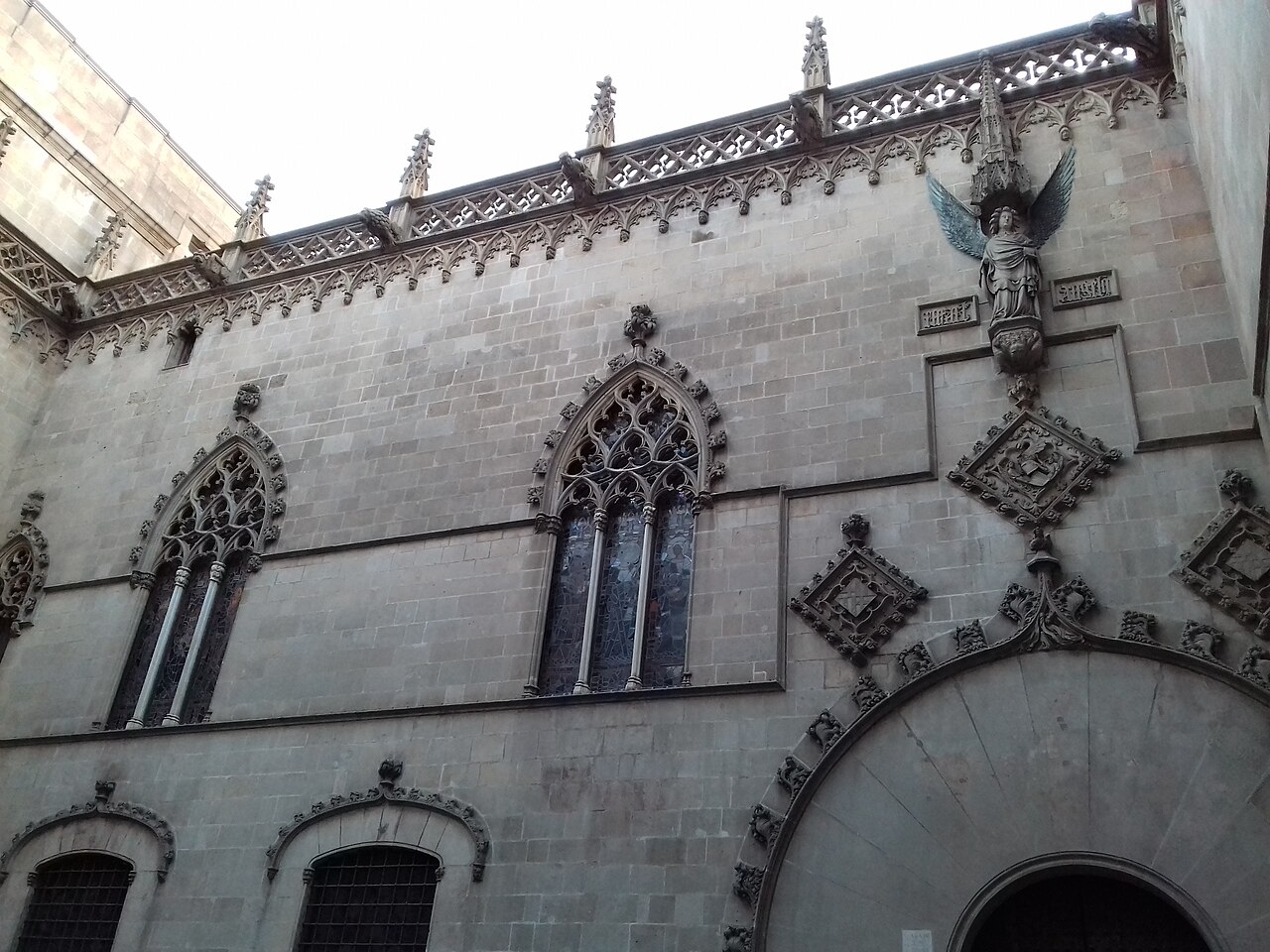 A stone wall with pointed arched windows and carved figures, seen from the street below.
