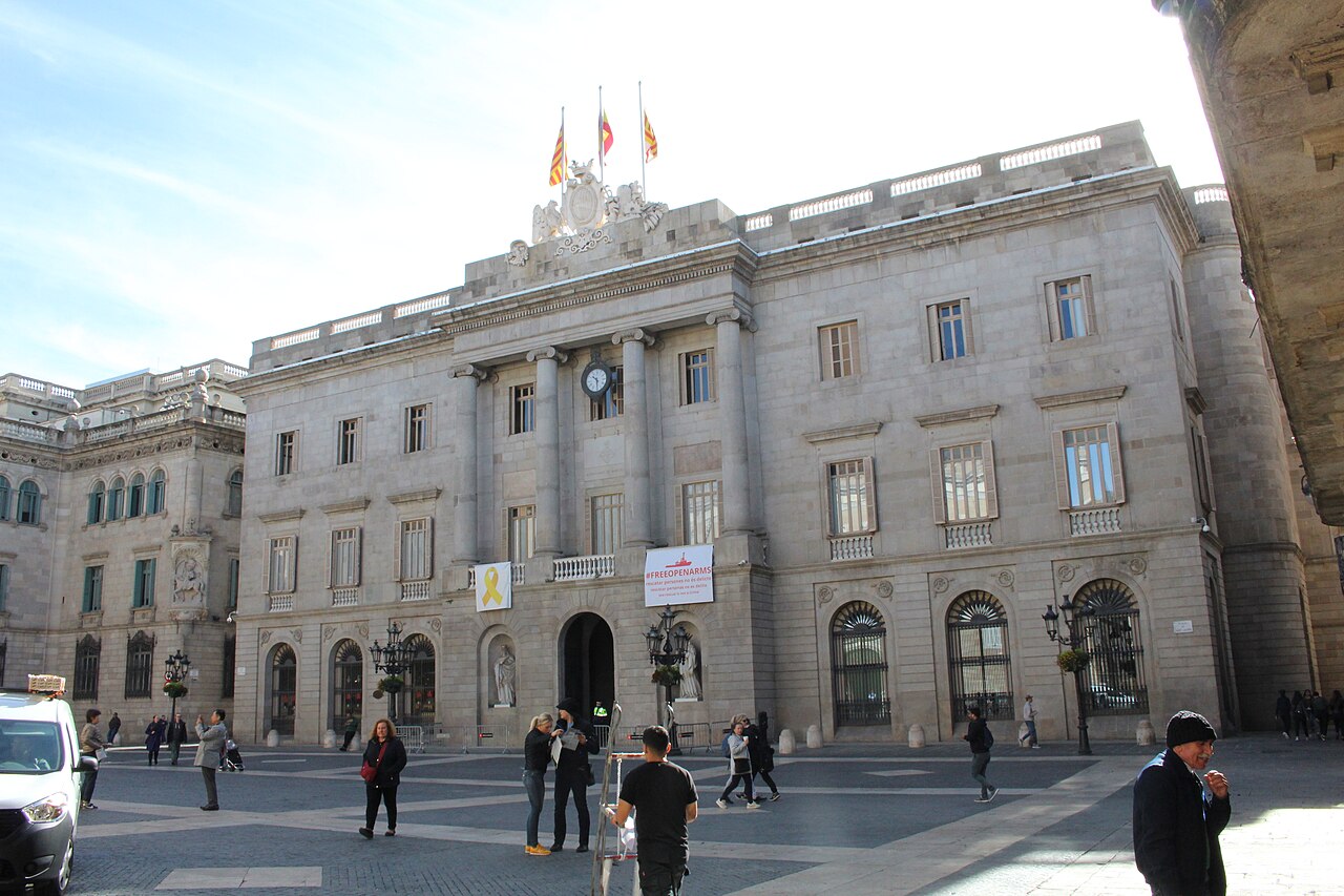 A grey neoclassical building with columns and flags faces a paved square with a few people crossing.
