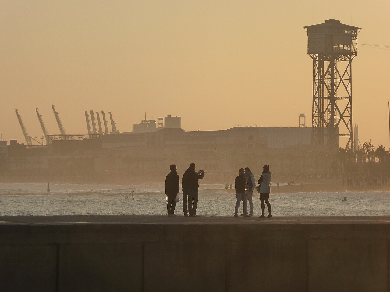 Small figures stand in silhouette on a beach under a hazy orange sky, beside a tall lattice tower.