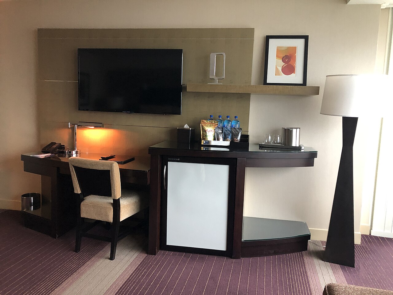 A dark wood desk and chair sit beside a cabinet with a small glass-fronted fridge, under a wall-mounted television.