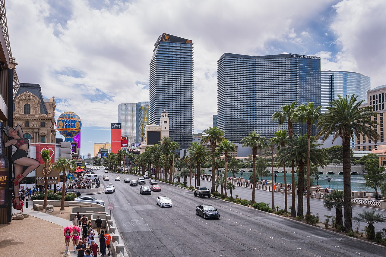 A wide boulevard lined with palm trees runs between tall glass hotel towers under a cloudy sky.