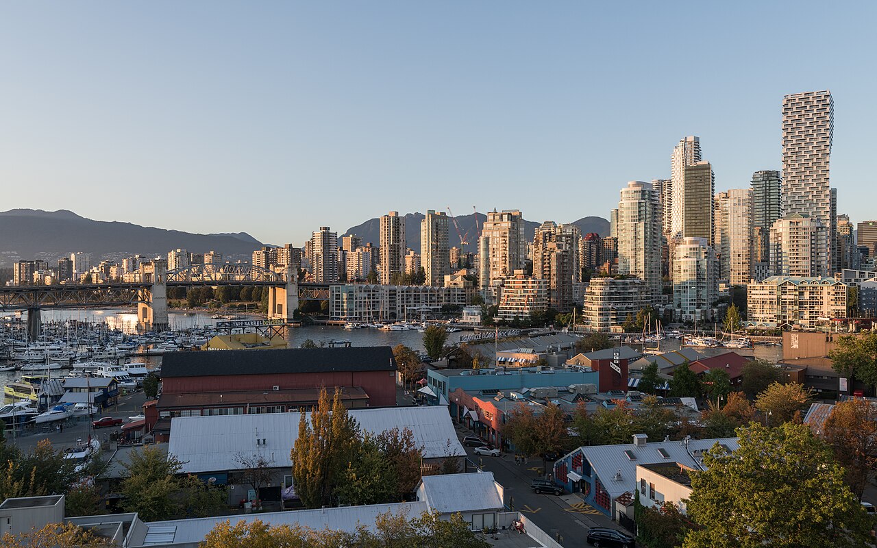 Warm evening light on a cluster of residential towers above a marina, low market buildings and a steel bridge, with mountains behind.