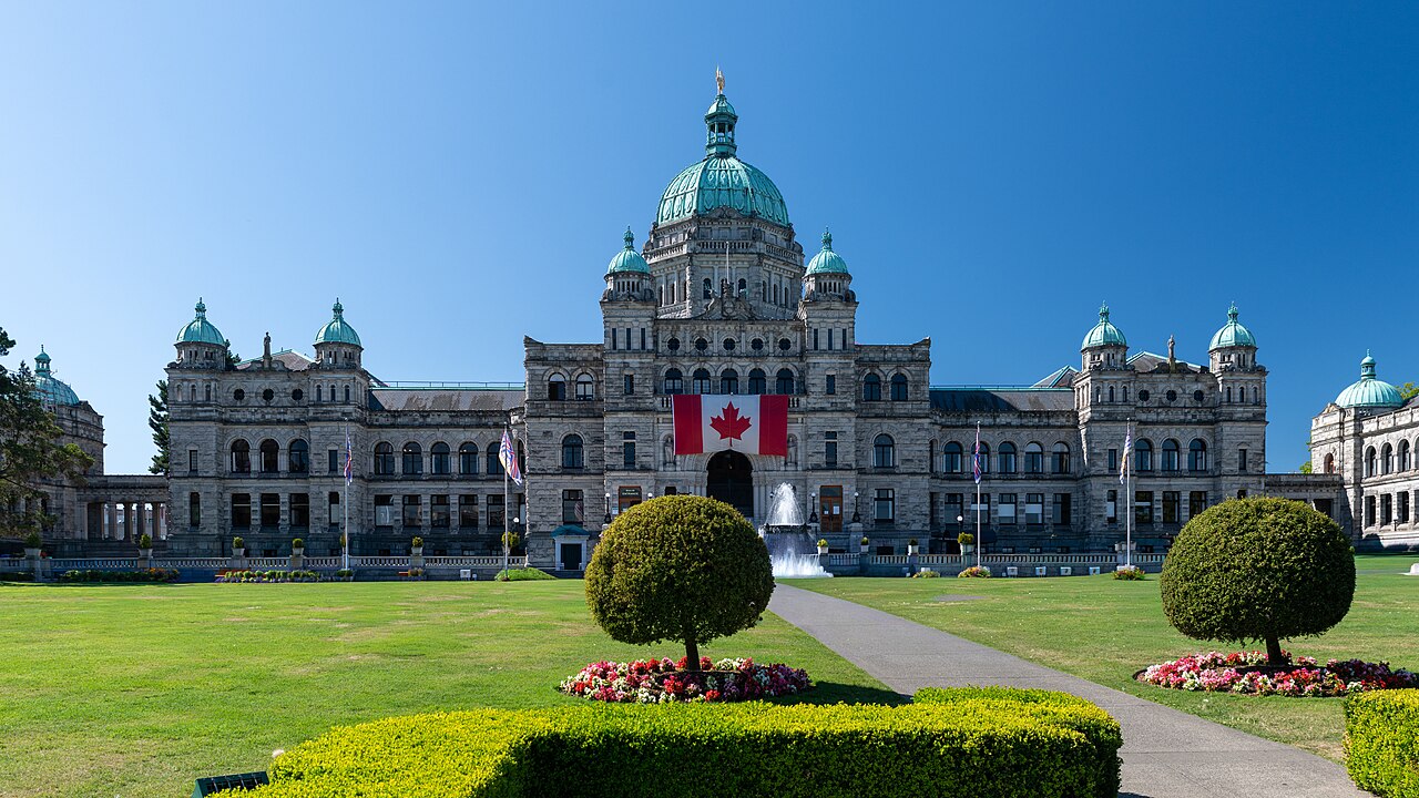 A grey stone building with green copper domes behind a lawn, clipped round shrubs and flower beds under a clear blue sky.