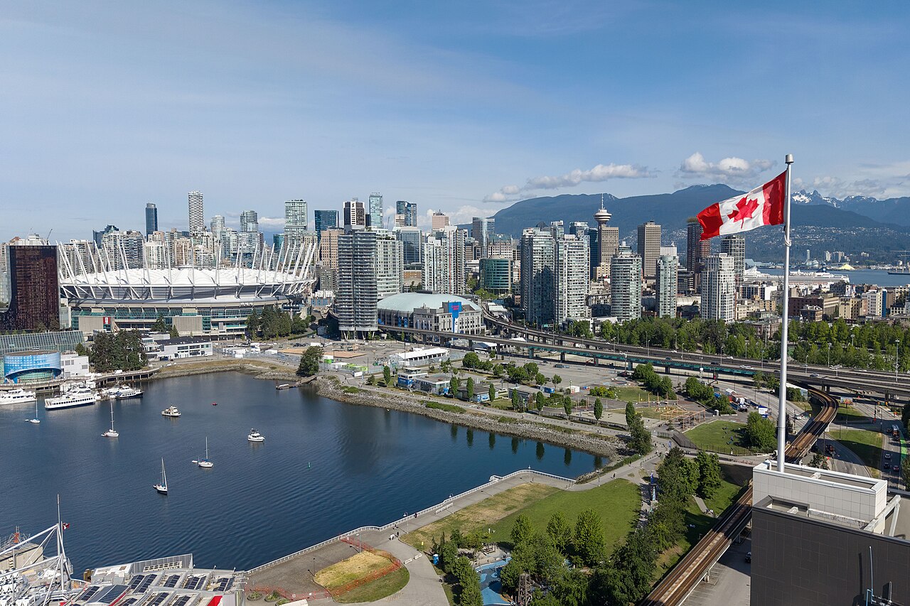 An inlet with moored boats and a white-roofed stadium in front of dense glass towers, a Canadian flag flying in the foreground.